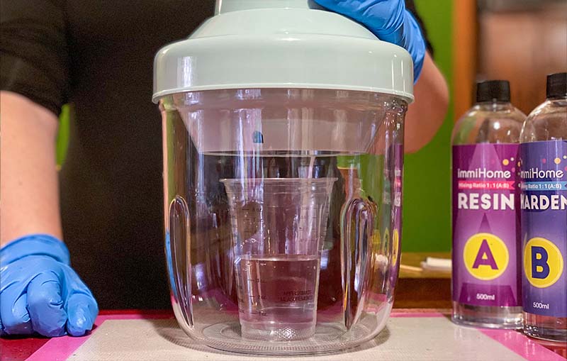 A person wearing blue gloves is using a vacuum chamber to degas a mixture for silicone molds for resin, with two bottles labeled 'Resin' and 'Hardener' placed nearby