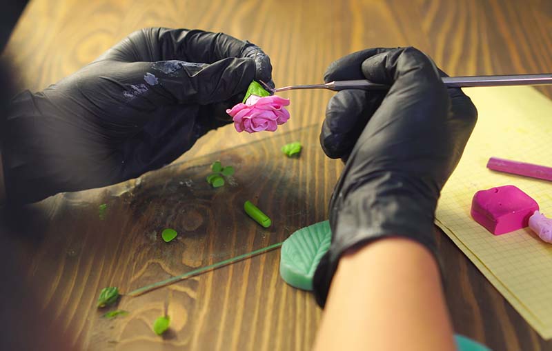 A close-up of an artist using sculpting tools to refine a delicate pink flower made from polymer clay molds. Black gloves and fine details highlight precision crafting.