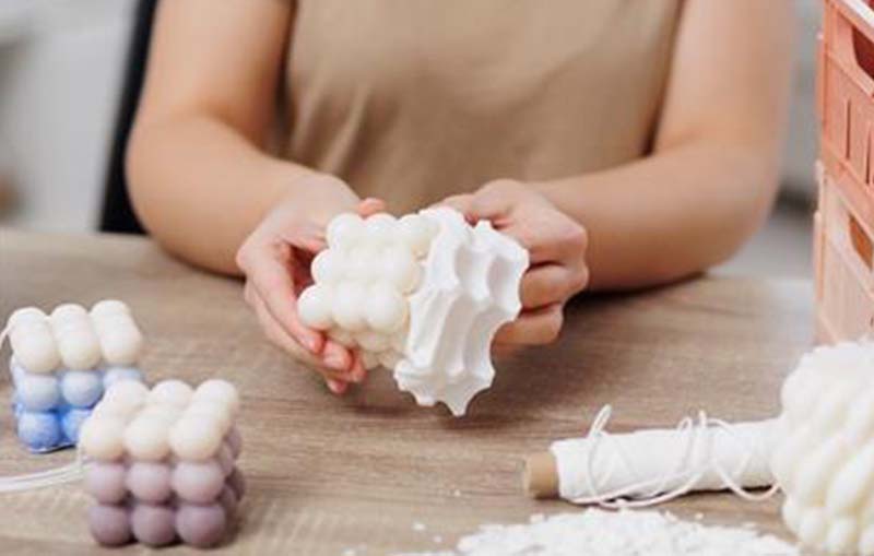 A woman carefully removes a bubble-shaped candle from a flexible silicone mold on a wooden craft table, with other molded candles nearby.