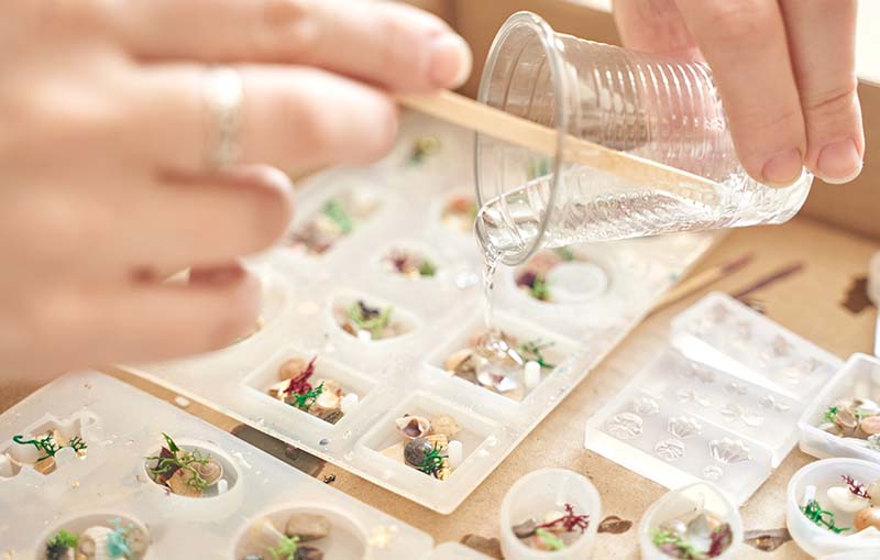 Close-up of hands pouring clear epoxy resin into small silicone molds filled with dried flowers and decorative elements for jewelry making.