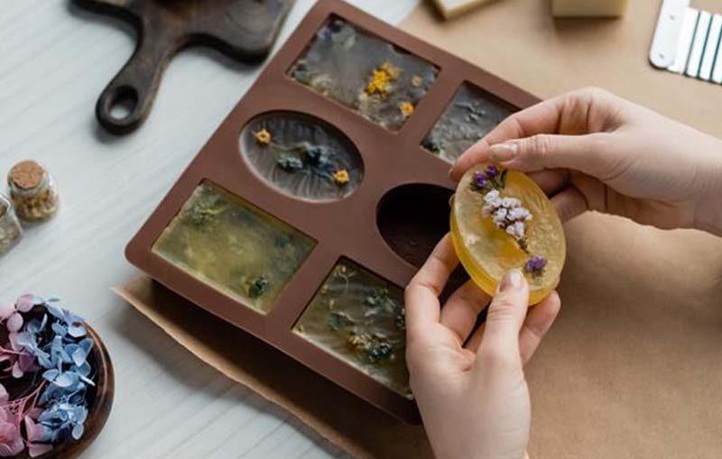 Hands carefully removing a handmade botanical soap infused with dried flowers from a brown silicone mold, with natural soap-making materials on the table.